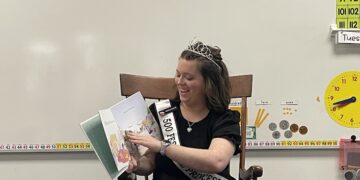 A smiling young woman, serving as a 500 Festival Princess for 2026, sits in a wooden chair in a classroom. She is wearing a black dress, a sparkling tiara, and her official white sash as she reads a colorful children's picture book aloud. The background features typical classroom decorations, including a number line, a chart of numbers up to 112, a yellow toy clock, and illustrations of coins and base-ten blocks.