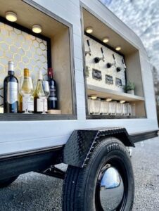 A close-up, low-angle shot of a white mobile bar trailer, focusing on its built-in beverage service areas. On the left, a recessed wooden shelf holds several wine bottles and glasses against a white and gold honeycomb-patterned backsplash. To the right, a larger service window features four beverage taps mounted on a white honeycomb wall, with a lower shelf holding rows of empty glasses. The trailer sits on a gravel surface, and a single black tire with a shiny chrome hubcap is prominent in the foreground.
