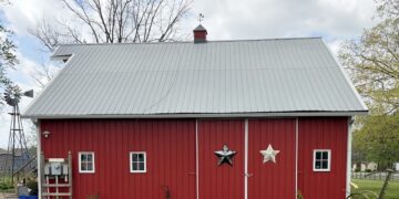 A classic red barn building with white trim around the windows and doors; a black and a white decorative star hang on the main sliding doors. A small windmill stands to the left against a backdrop of bare trees and a cloudy sky.