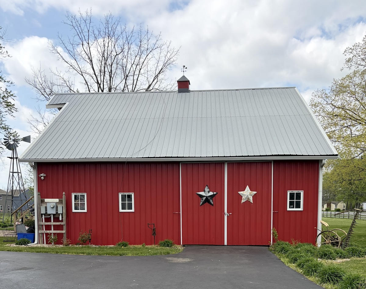 A classic red barn building with white trim around the windows and doors; a black and a white decorative star hang on the main sliding doors. A small windmill stands to the left against a backdrop of bare trees and a cloudy sky.