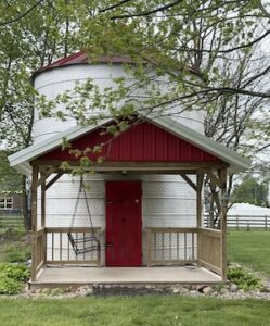 A repurposed farm silo with a red metal roof and matching red door; it has a timber-frame front porch with a gabled roof, set in a green backyard with leafy trees.