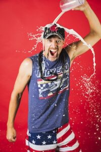 A man with a mustache and an enthusiastic expression stands against a vibrant red background. He is wearing a dark blue Indy 500 sleeveless t-shirt, a matching baseball cap, and American flag patterned shorts. He holds a clear container above his head, pouring a stream of milk that splashes over his hat and down his face. A black racing-style stethoscope or cable is draped around his neck. The man’s mouth is open in a shout, and his right fist is clenched in a pose of victory.