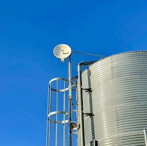 A white parabolic dish antenna is mounted to a metal pole on the side of a tall, corrugated silver grain silo. A safety ladder with a protective cage is attached to the silo next to the antenna. The entire scene is set against a clear, solid blue sky.