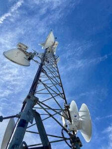A low-angle shot looking up a tall, lattice-style telecommunications tower against a bright blue sky with wispy clouds. Multiple white parabolic dish antennas are mounted at various heights along the metal structure, pointing in different directions.