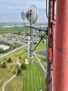 A high-angle view from a telecommunications tower looking down at a golf course and a residential neighborhood. In the foreground, a white parabolic dish antenna and a vertical sector antenna are mounted on a gray metal pole attached to a red tower leg. The background shows green fairways, sand traps, a small pond, and rows of suburban houses.