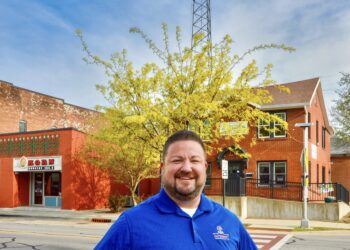 A smiling man in a bright blue polo shirt with a company logo stands on a sidewalk. He has his hands on his hips. Behind him are two brick buildings, a vibrant yellow tree, and a very tall communication tower against a clear blue sky. On the left, a red brick building features a sign for KORN Country 100.3.