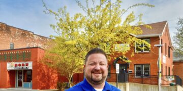 A smiling man in a bright blue polo shirt with a company logo stands on a sidewalk. He has his hands on his hips. Behind him are two brick buildings, a vibrant yellow tree, and a very tall communication tower against a clear blue sky. On the left, a red brick building features a sign for KORN Country 100.3.