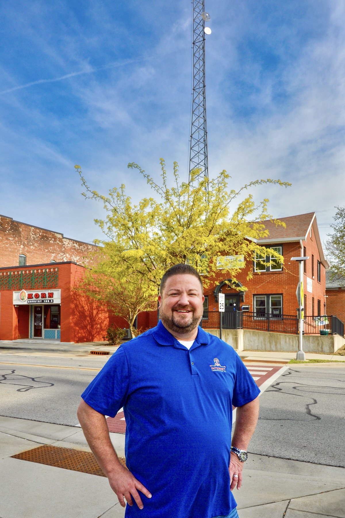 A smiling man in a bright blue polo shirt with a company logo stands on a sidewalk. He has his hands on his hips. Behind him are two brick buildings, a vibrant yellow tree, and a very tall communication tower against a clear blue sky. On the left, a red brick building features a sign for KORN Country 100.3.
