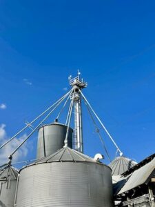 A wide shot of several corrugated silver grain bins at a farm. In the center, a tall grain elevator tower rises high above the bins, topped with a metal platform. Small wireless internet antennas are visible on top of the tower's platform, reaching toward a clear, deep blue sky.