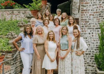 A group of fourteen diverse women of various ages smiling and posing together on outdoor brick stairs. They are dressed in business-casual attire in soft, neutral, and pastel tones. The background features lush greenery and classic brick architecture.