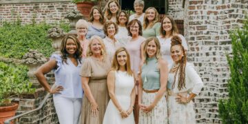 A group of fourteen diverse women of various ages smiling and posing together on outdoor brick stairs. They are dressed in business-casual attire in soft, neutral, and pastel tones. The background features lush greenery and classic brick architecture.