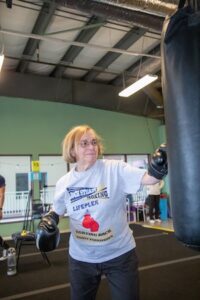 A woman with blonde hair and glasses is captured in a boxing gym, throwing a punch at a large black heavy bag. She is wearing black boxing gloves and a light grey t-shirt that reads "Rock Steady Boxing Lifeplex" with the tagline "Fighting Back Against Parkinson's." The gym has high ceilings with exposed industrial pipes and green walls in the background.