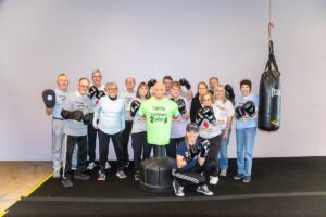 A large group of smiling seniors and a trainer pose together in a boxing gym. Most are wearing boxing gloves and t-shirts that say "Rock Steady Boxing." In the center of the group stands a green torso-shaped punching dummy wearing a shirt that reads "Fighting Parkinson’s 2gether." A heavy Everlast punching bag hangs to the right, and the group is gathered on a black gym mat against a plain grey wall.