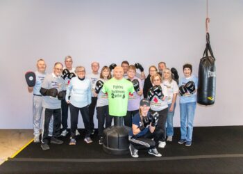 A large group of smiling seniors and a trainer pose together in a boxing gym. Most are wearing boxing gloves and t-shirts that say "Rock Steady Boxing." In the center of the group stands a green torso-shaped punching dummy wearing a shirt that reads "Fighting Parkinson’s 2gether." A heavy Everlast punching bag hangs to the right, and the group is gathered on a black gym mat against a plain grey wall.