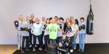 A large group of smiling seniors and a trainer pose together in a boxing gym. Most are wearing boxing gloves and t-shirts that say "Rock Steady Boxing." In the center of the group stands a green torso-shaped punching dummy wearing a shirt that reads "Fighting Parkinson’s 2gether." A heavy Everlast punching bag hangs to the right, and the group is gathered on a black gym mat against a plain grey wall.