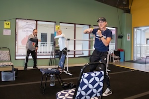 An indoor fitness class at the Lifeplex facility, where a female instructor in a navy blue t-shirt and baseball cap is leading a session. She is demonstrating a boxing-style punch for two senior participants who are following along in the background. The gym features green and yellow walls, large windows, and black mats. A chair with a blue and white patterned cushion is in the foreground, part of a circuit-style workout setup.