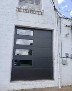 A vertical shot of a contemporary Skyline Flush style garage door in a dark finish, featuring four rectangular glass windows that reflect the sky, set against a white masonry exterior.