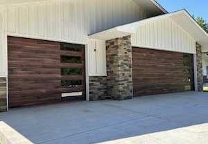 A low-angle outdoor shot of a contemporary garage featuring dark woodgrain plank doors with narrow vertical windows on the right side, complemented by a white board-and-batten exterior and grey stonework.