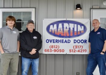 Three men representing Martin Overhead Door stand in front of a grey building with a large banner displaying the company logo and contact phone numbers for Greensburg (812-663-9050) and the surrounding area (317-512-4219).