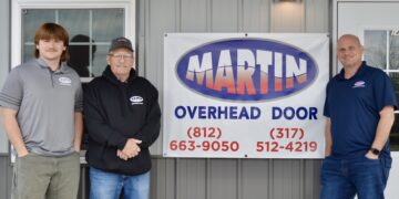 Three men representing Martin Overhead Door stand in front of a grey building with a large banner displaying the company logo and contact phone numbers for Greensburg (812-663-9050) and the surrounding area (317-512-4219).