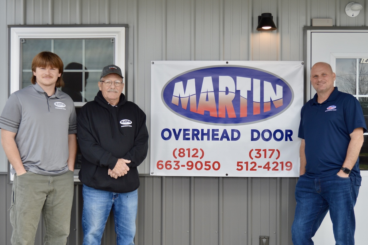 Three men representing Martin Overhead Door stand in front of a grey building with a large banner displaying the company logo and contact phone numbers for Greensburg (812-663-9050) and the surrounding area (317-512-4219).