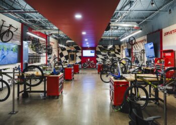 A wide, brightly lit bicycle repair workshop with polished concrete floors. A red ceiling strip runs down the center of the room, leading toward a black-and-white checkered wall. Multiple wooden workbenches with red tool chests are arranged on both sides, each equipped with a bike repair stand. Bicycles in various stages of assembly are mounted on the stands or lean against the walls. Large signs on the right wall read "DRIVETRAIN" and "COMPONENTS" in bold red lettering.
