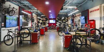 A wide, brightly lit bicycle repair workshop with polished concrete floors. A red ceiling strip runs down the center of the room, leading toward a black-and-white checkered wall. Multiple wooden workbenches with red tool chests are arranged on both sides, each equipped with a bike repair stand. Bicycles in various stages of assembly are mounted on the stands or lean against the walls. Large signs on the right wall read "DRIVETRAIN" and "COMPONENTS" in bold red lettering.