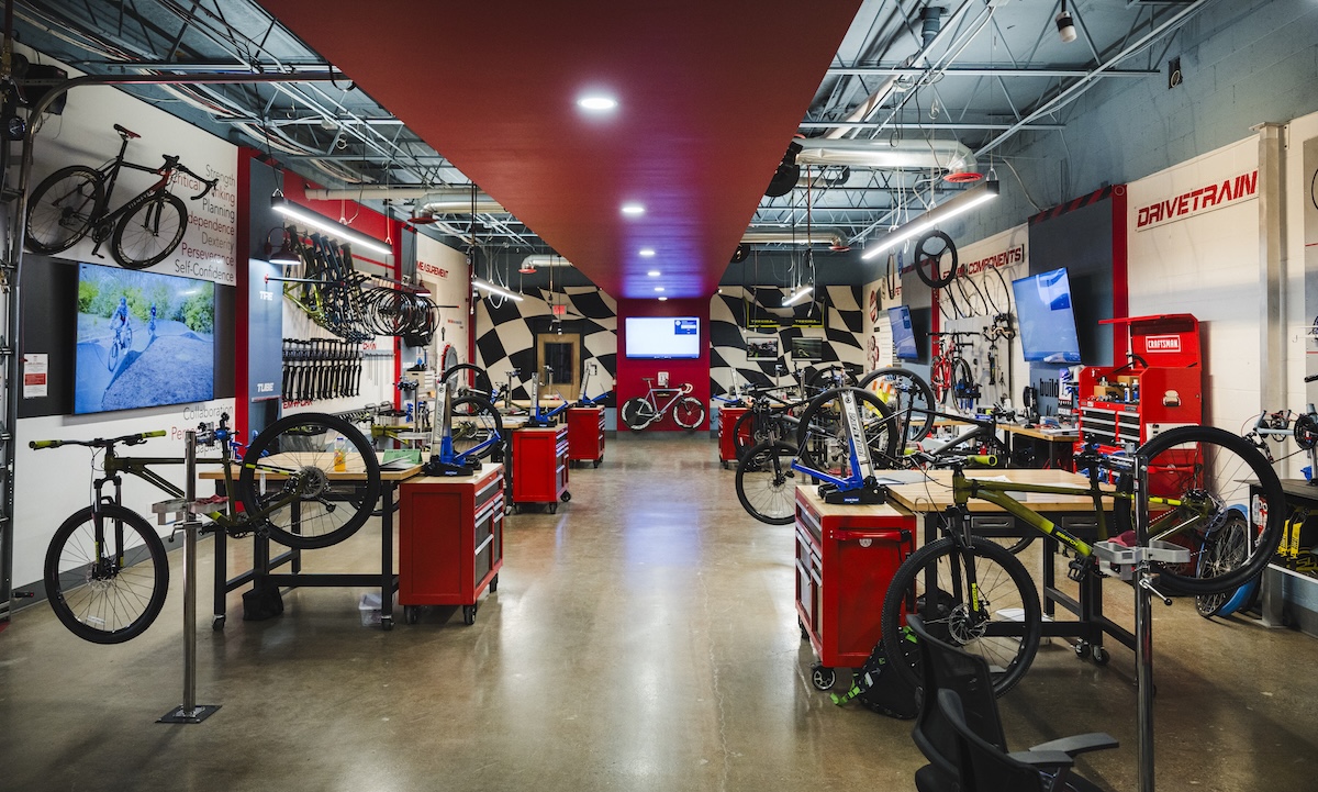 A wide, brightly lit bicycle repair workshop with polished concrete floors. A red ceiling strip runs down the center of the room, leading toward a black-and-white checkered wall. Multiple wooden workbenches with red tool chests are arranged on both sides, each equipped with a bike repair stand. Bicycles in various stages of assembly are mounted on the stands or lean against the walls. Large signs on the right wall read "DRIVETRAIN" and "COMPONENTS" in bold red lettering.