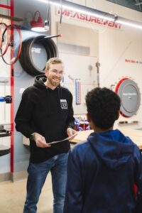 A smiling man with glasses, wearing a black Nine13sports hoodie, talks to a young boy in a workshop. The man holds a piece of paper and stands in front of a wall with "MEASUREMENT" written on it. Various bicycle parts, including a tire and wheels, are mounted on the wall behind them. The boy is seen from behind, wearing a navy blue hoodie, listening to the instructor in a brightly lit community center setting.