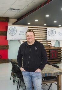 A smiling man with light brown hair and a beard stands in a modern community room, leaning slightly against a wooden table. He is wearing a black Nine13sports zip-up hoodie over a black shirt and light-wash blue jeans. In the background, there is a wood-slat wall with monitors displaying "Runner Bundas" and a stylized compass logo. The room is brightly lit with recessed ceiling lights and features red and grey architectural accents.
