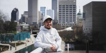 A smiling man wearing a white baseball cap and a light grey hoodie leans against a white railing on a bridge. The hoodie features a small Nike logo and Korean text that reads "그냥 했어" (Just Do It). In the background, the downtown Indianapolis skyline stands tall under a bright sky, with the Central Canal visible below.