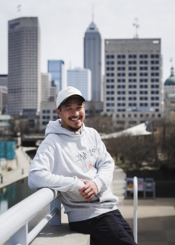 A smiling man wearing a white baseball cap and a light grey hoodie leans against a white railing on a bridge. The hoodie features a small Nike logo and Korean text that reads "그냥 했어" (Just Do It). In the background, the downtown Indianapolis skyline stands tall under a bright sky, with the Central Canal visible below.