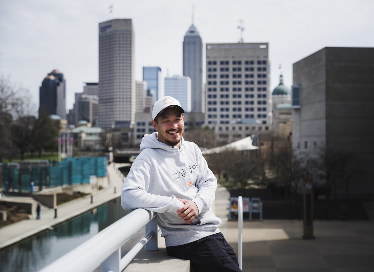 A smiling man wearing a white baseball cap and a light grey hoodie leans against a white railing on a bridge. The hoodie features a small Nike logo and Korean text that reads "그냥 했어" (Just Do It). In the background, the downtown Indianapolis skyline stands tall under a bright sky, with the Central Canal visible below.