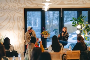 In a room with an artistic, mural-covered wall, a man and a woman sit at the front, engaged in a discussion or interview. The woman, wearing a black leather jacket, speaks into a microphone while the man, in a tan jacket and white beanie, looks toward her. An audience of several people is seated in the foreground, with one person holding up a smartphone to record or take a photo of the speakers. Large windows in the background reveal a soft, outdoor light.