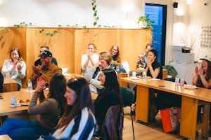 A group of about ten people are seated around long wooden tables in a cozy, modern room with light wood paneled walls decorated with hanging plants. Most of the people are smiling and clapping their hands, looking toward something off-camera. The atmosphere is warm and celebratory, with soft indoor lighting and some food and drinks visible on the tables.
