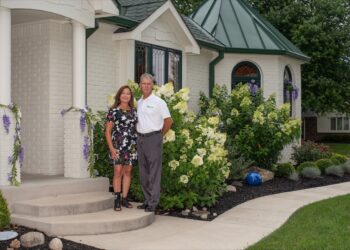 A full-shot of a man and woman standing together in front of a white brick house with a distinctive green metal-roofed turret. The man is wearing a white polo shirt and grey trousers, while the woman wears a black, short-sleeved mini dress with a vibrant white, yellow, and pink floral pattern. They are framed by lush landscaping, including massive, blooming white hydrangeas, purple wisteria vines climbing a porch pillar, and a dark-mulched garden bed with a single blue decorative glass ball. A curved concrete walkway leads to the front steps.