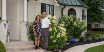 A full-shot of a man and woman standing together in front of a white brick house with a distinctive green metal-roofed turret. The man is wearing a white polo shirt and grey trousers, while the woman wears a black, short-sleeved mini dress with a vibrant white, yellow, and pink floral pattern. They are framed by lush landscaping, including massive, blooming white hydrangeas, purple wisteria vines climbing a porch pillar, and a dark-mulched garden bed with a single blue decorative glass ball. A curved concrete walkway leads to the front steps.