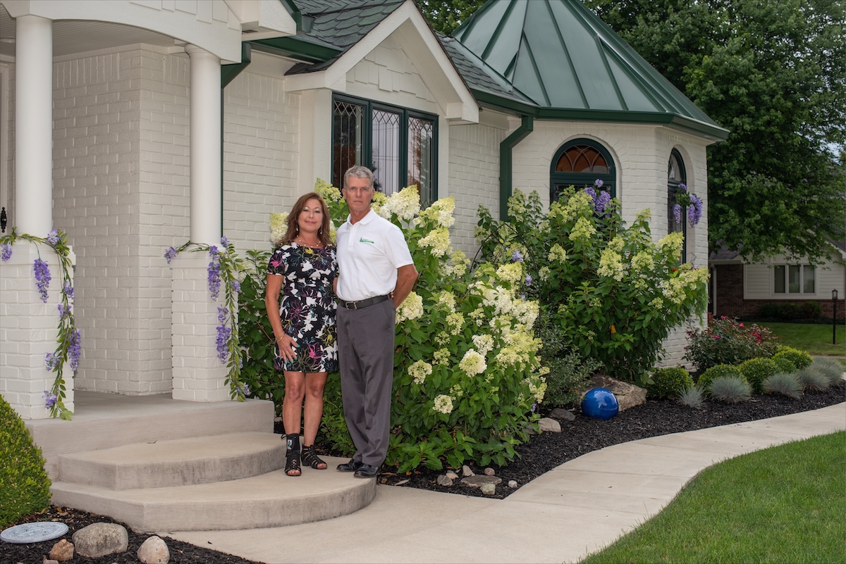 A full-shot of a man and woman standing together in front of a white brick house with a distinctive green metal-roofed turret. The man is wearing a white polo shirt and grey trousers, while the woman wears a black, short-sleeved mini dress with a vibrant white, yellow, and pink floral pattern. They are framed by lush landscaping, including massive, blooming white hydrangeas, purple wisteria vines climbing a porch pillar, and a dark-mulched garden bed with a single blue decorative glass ball. A curved concrete walkway leads to the front steps.