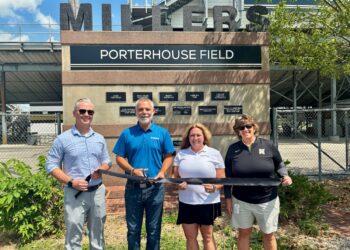 A bright outdoor shot of a ribbon-cutting ceremony at PorterHouse Contracting Field in Noblesville, Indiana. Standing in front of a tan stone and brick monument that reads "MILLERS - PORTERHOUSE FIELD," four individuals hold a long black ribbon. From left to right are Noblesville Schools Superintendent Dr. Dan Hile, James Porter, Kristi Porter (owners of PorterHouse Contracting), and Noblesville High School Athletic Director Leah Wooldridge. The monument features several black plaques recognizing various sponsors. In the background, the metal bleachers and fencing of the sports complex are visible under a clear, sunny sky.