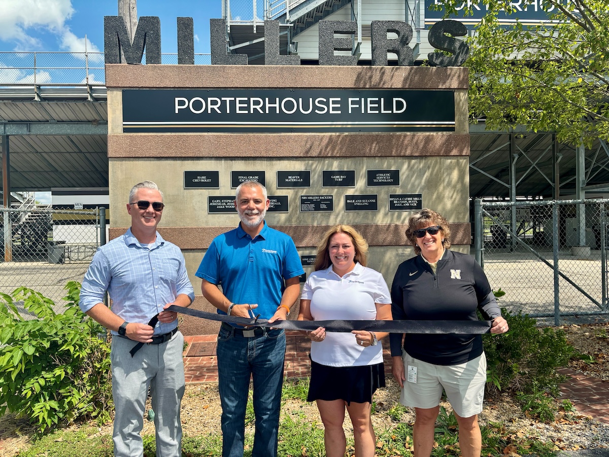 A bright outdoor shot of a ribbon-cutting ceremony at PorterHouse Contracting Field in Noblesville, Indiana. Standing in front of a tan stone and brick monument that reads "MILLERS - PORTERHOUSE FIELD," four individuals hold a long black ribbon. From left to right are Noblesville Schools Superintendent Dr. Dan Hile, James Porter, Kristi Porter (owners of PorterHouse Contracting), and Noblesville High School Athletic Director Leah Wooldridge. The monument features several black plaques recognizing various sponsors. In the background, the metal bleachers and fencing of the sports complex are visible under a clear, sunny sky.