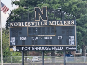 A detailed view of the large, high-school-style scoreboard at PorterHouse Contracting Field in Noblesville, Indiana. The scoreboard features a black background with white and yellow lettering. At the top, a prominent black-and-gold "N" logo is centered within a decorative arch, flanked by the words "NOBLESVILLE MILLERS". Below the digital clock and score displays, a large black-and-white sign reads "PORTERHOUSE FIELD". The bottom right of this sign includes the blue-and-gray logo for PorterHouse Contracting. The scoreboard is set against a background of mature green trees, with an American flag visible on a pole to the left.