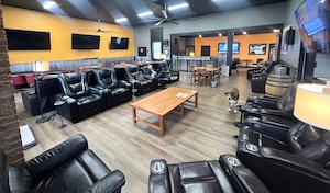 The wide-angle interior shot reveals the spacious main lounge of the Cigar Club of Indiana in Greenwood. Rows of plush black leather recliners with built-in cup holders face each other, separated by a large, rectangular wooden coffee table in the center. The room features warm wood-look flooring, light grey walls with corrugated metal wainscoting, and a bold yellow accent wall adorned with three large flat-screen TVs. Contemporary lighting, including floor lamps and ceiling fans, illuminates the space, while a glimpse of a smaller seating area and a humidor can be seen in the background.