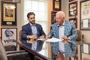 Two men in business attire sit at a dark, reflective conference table, focused on a document they are reviewing together. The younger man on the left wears a navy suit jacket over a light blue shirt, while the older man on the right wears a blue and grey plaid blazer. They are surrounded by framed memorabilia on the walls, including a notable "VOTE" poster featuring Barack Obama.