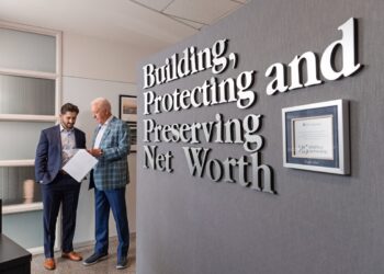 Two men in professional attire review documents in a modern office. On the right, a grey wall features large 3D silver letters that read, "Building, Protecting and Preserving Net Worth." Next to the text hangs a framed "25 years of partnership" certificate from LPL Financial. The man on the left wears a navy suit, and the older man on the right wears a blue plaid blazer.