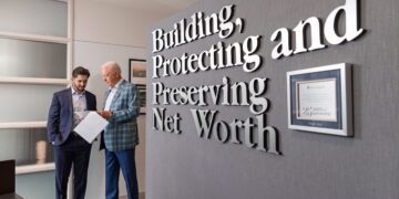 Two men in professional attire review documents in a modern office. On the right, a grey wall features large 3D silver letters that read, "Building, Protecting and Preserving Net Worth." Next to the text hangs a framed "25 years of partnership" certificate from LPL Financial. The man on the left wears a navy suit, and the older man on the right wears a blue plaid blazer.