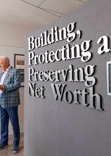 Two men in professional attire review documents in a modern office. On the right, a grey wall features large 3D silver letters that read, "Building, Protecting and Preserving Net Worth." Next to the text hangs a framed "25 years of partnership" certificate from LPL Financial. The man on the left wears a navy suit, and the older man on the right wears a blue plaid blazer.