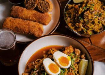 A wooden table features an overhead spread of New Orleans-style Creole-Asian fusion cuisine, including a large bowl of Yakamein noodles topped with a soft-boiled egg cut in half, sliced green onions, and a lime wedge. Accompanying dishes include a plate of golden fried egg rolls sliced open, a small metal bowl filled with seasoned fried rice, a tall glass of soda, and a glass of beer.