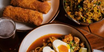 A wooden table features an overhead spread of New Orleans-style Creole-Asian fusion cuisine, including a large bowl of Yakamein noodles topped with a soft-boiled egg cut in half, sliced green onions, and a lime wedge. Accompanying dishes include a plate of golden fried egg rolls sliced open, a small metal bowl filled with seasoned fried rice, a tall glass of soda, and a glass of beer.