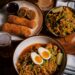 A wooden table features an overhead spread of New Orleans-style Creole-Asian fusion cuisine, including a large bowl of Yakamein noodles topped with a soft-boiled egg cut in half, sliced green onions, and a lime wedge. Accompanying dishes include a plate of golden fried egg rolls sliced open, a small metal bowl filled with seasoned fried rice, a tall glass of soda, and a glass of beer.