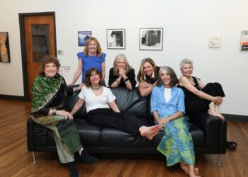 Seven women pose together around a black leather couch inside an art gallery with white walls and hardwood floors. Framed artwork is hanging on the wall behind them, and a wooden door with the number "212" is visible in the background.